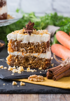 Een stuk zelfgebakken carrot cake met glazuur. Wortels op de achtergrond en kaneel op de voorgrond